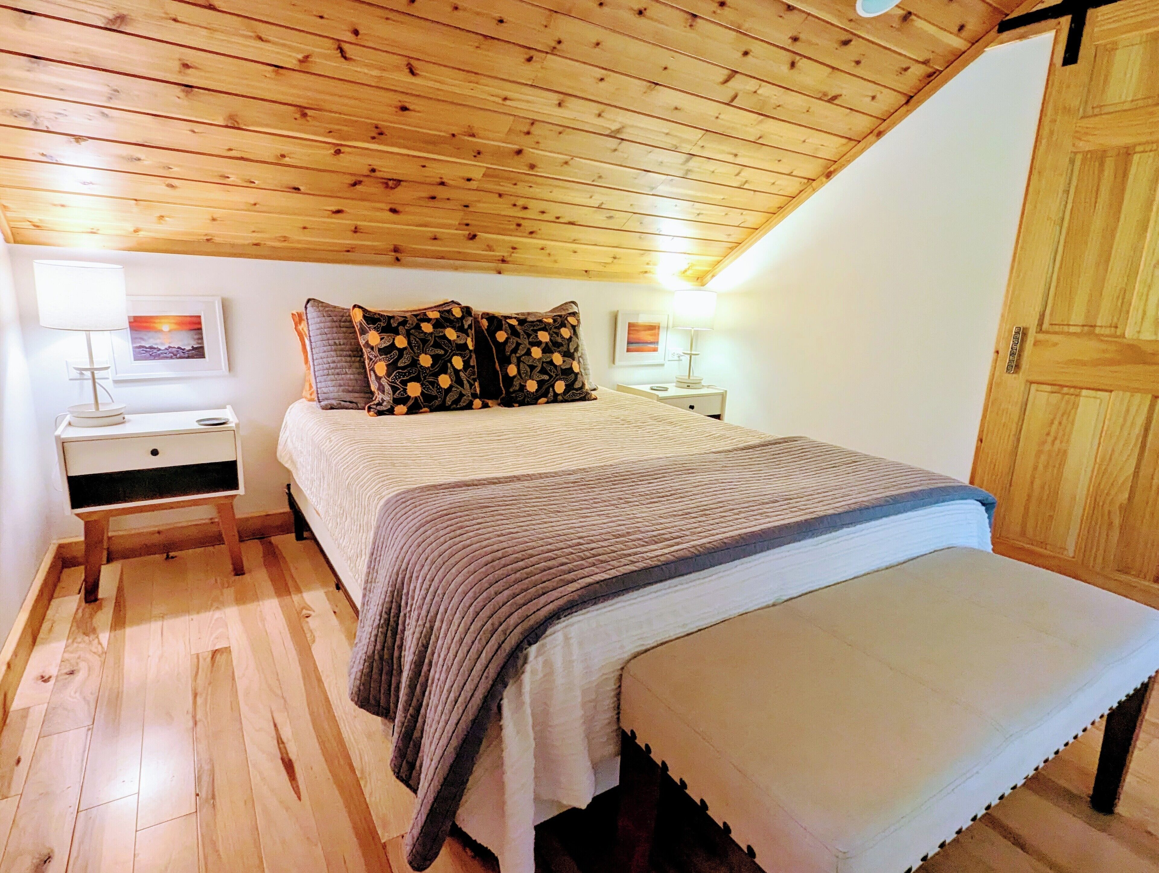 Un dormitorio con techo de madera, una cama con colcha gris, dos mesitas de noche con lámparas y una puerta de madera.