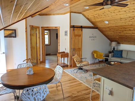 A cozy, well-lit room with wooden flooring, a dining area with a round table and white chairs, a sofa, and a ceiling fan.