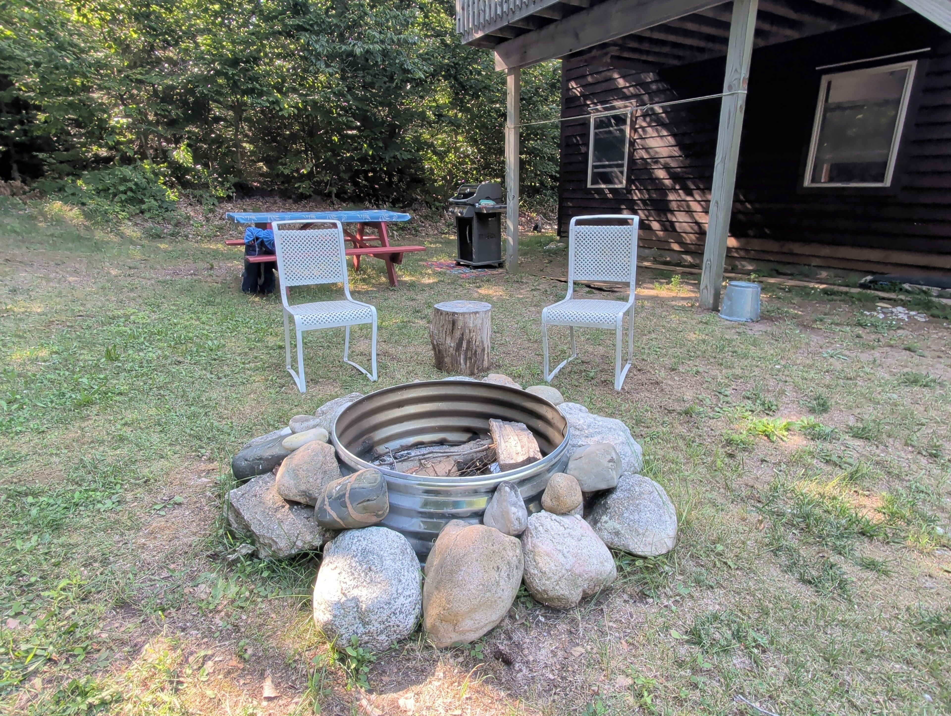 Een buitenzitplaats met een vuurkorf omringd door rotsen, twee witte metalen stoelen en een picknicktafel in een gazon met een houten hutje op de achtergrond.