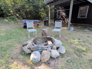 An outdoor seating area with a fire pit surrounded by rocks, two white metal chairs, and a picnic table in a grassy backyard with a wooden cabin in the background.