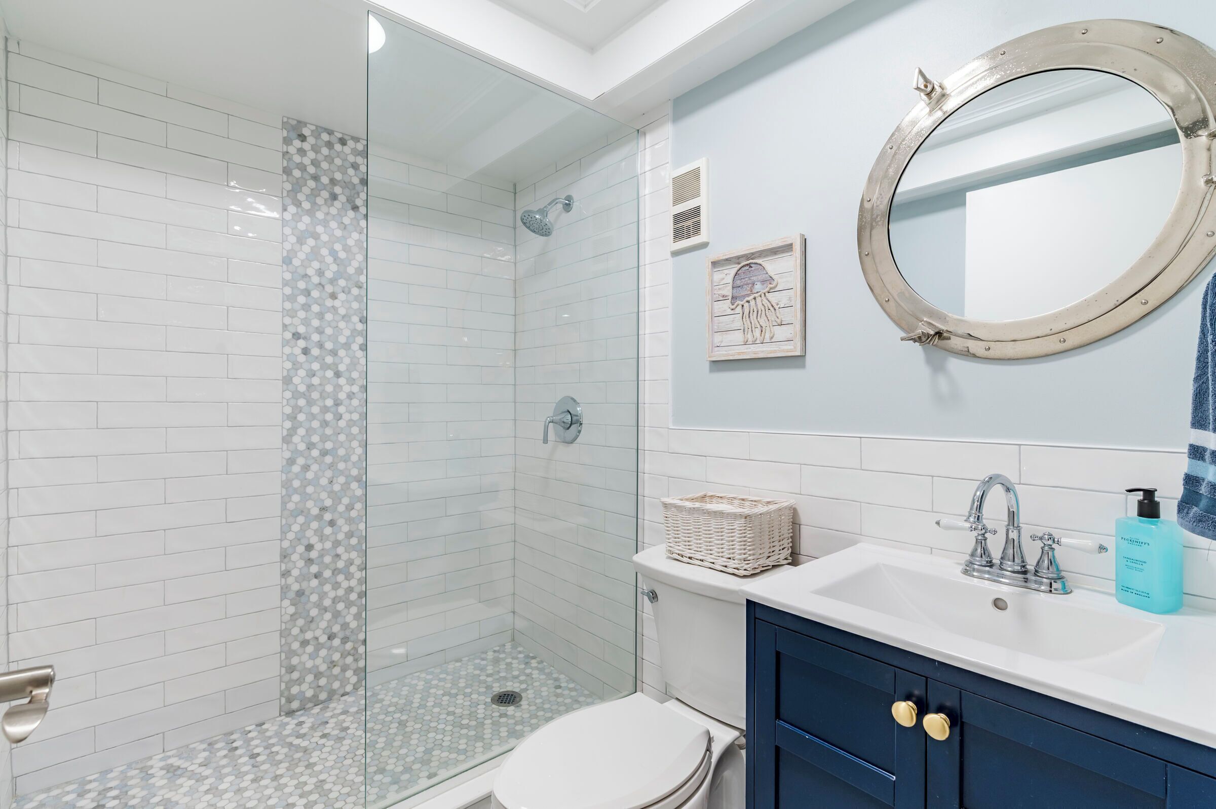 A modern bathroom with a glass-enclosed shower, a white toilet, a sink with a blue soap dispenser, and a decorative mirror.