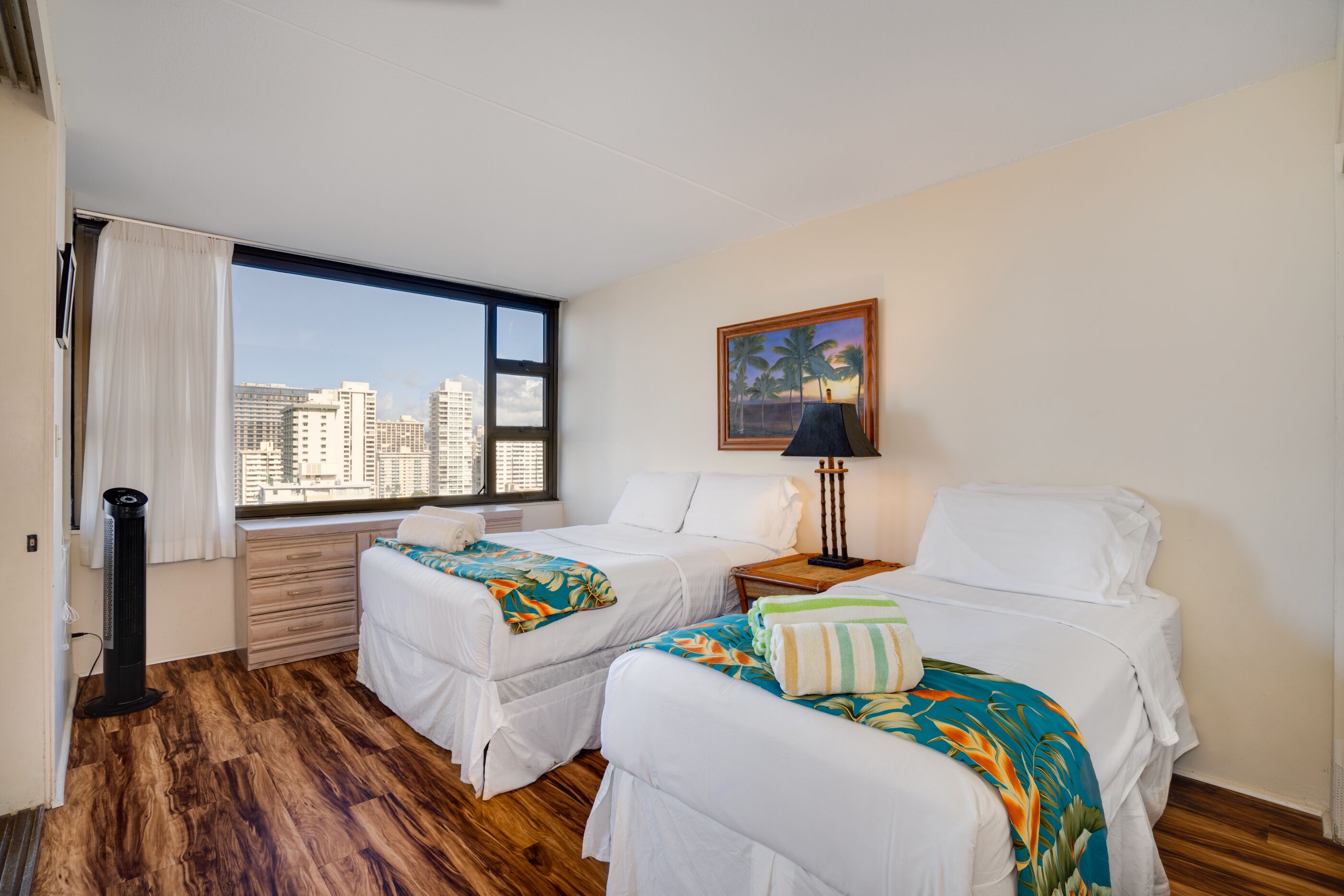 A hotel room with two beds, a large window with a city view, a standing heater, and a painting on the wall.