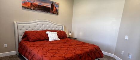 Un dormitorio con una cama grande, cabecera, mesita de noche, lámpara y un cuadro en la pared.