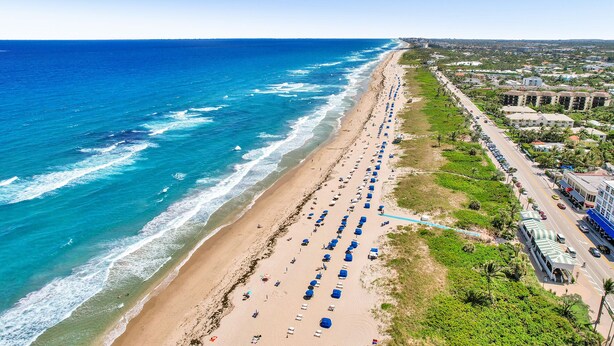 Ein Luftbild eines Strandes mit Reihen von Strandsonnenstühlen und Sonnenschirmen, einer parallel zum Strand verlaufenden Straße und Gebäuden im Hintergrund.
