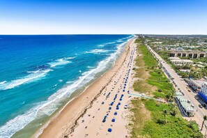 Ein Luftbild eines Strandes mit Reihen von Strandsonnenstühlen und Sonnenschirmen, einer parallel zum Strand verlaufenden Straße und Gebäuden im Hintergrund.