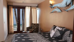Un dormitorio con cama, una silla, vista al océano a través de una puerta de vidrio y una escultura de delfín en la pared.