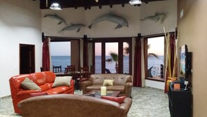 Sala de estar con dos sofás de cuero, una mesa de centro y un televisor. Amplias ventanas con cortinas, vista al océano y murales de delfines en el techo.