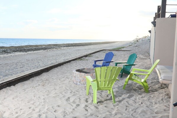 Una playa con tres sillas Adirondack, una pequeña mesa y un camino de hormigón que lleva al agua.