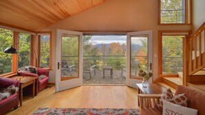 A cozy room with a wooden ceiling, a large sliding glass door leading to an outdoor deck, a plush red sofa, a small wooden table, and a view of autumn trees.