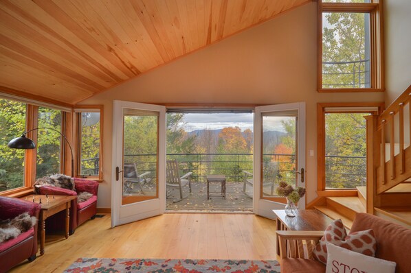 A cozy room with a wooden ceiling, a large sliding glass door leading to an outdoor deck, a plush red sofa, a small wooden table, and a view of autumn trees.