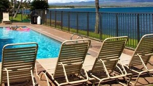 Zona de piscina con sillas de descanso y palmeras, con vista a un cuerpo de agua y tierra distante.