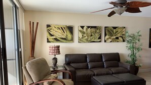 Una sala de estar con un sofá de cuero marrón, un sillón beige, un reposapiés negro y una lámpara de pie. Hay tres cuadros de plantas colgados en la pared y un ventilador de techo.