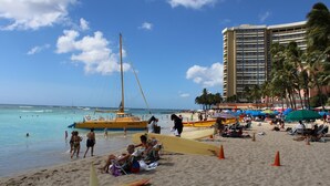 A beach with a tall building, palm trees, and a sailboat.