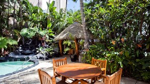 A wooden table and chairs set outdoors with a thatched-roof structure, surrounded by lush greenery and a small waterfall.