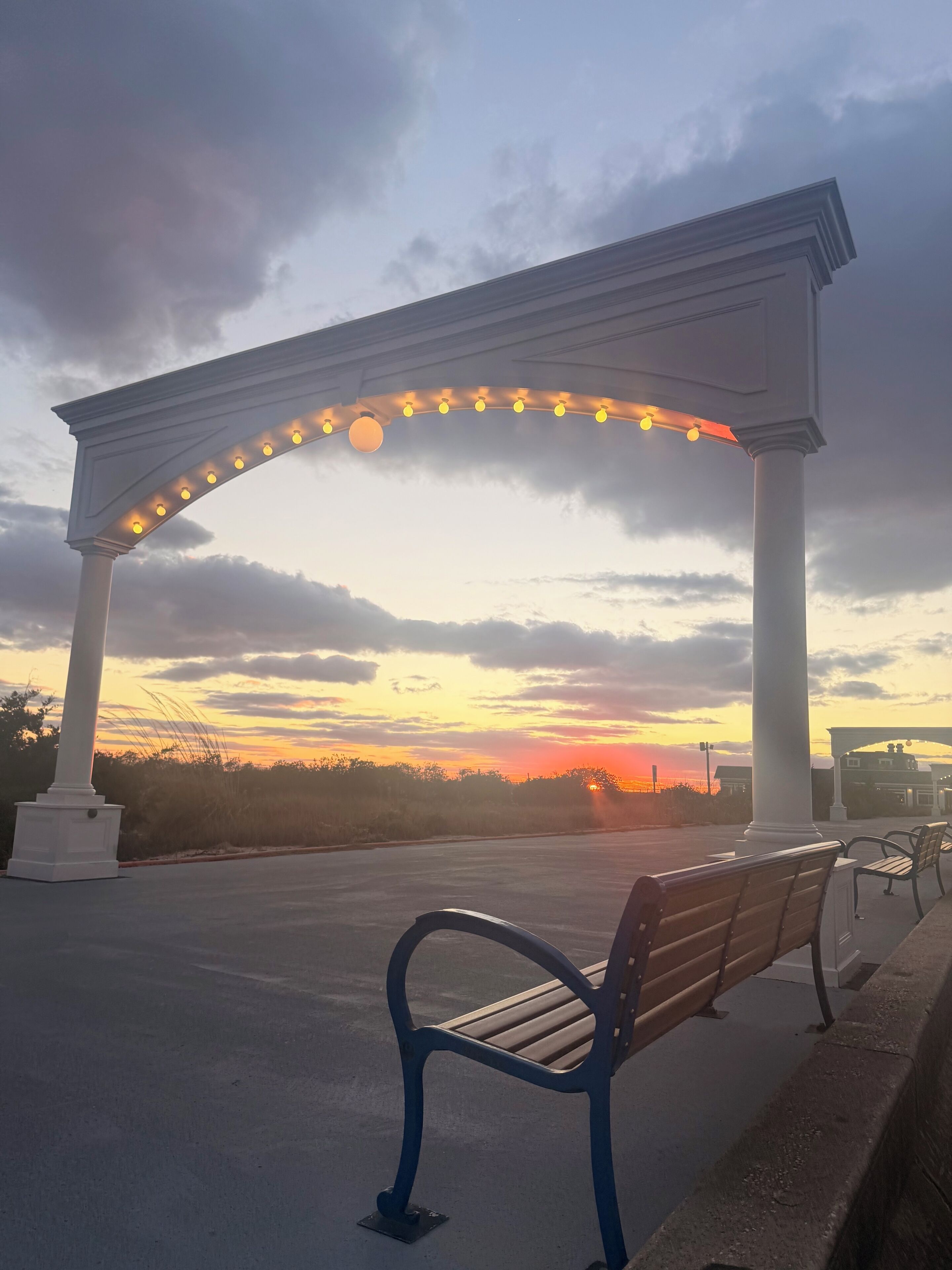 Un banco debajo de un arco blanco con luces, con vistas a la puesta de sol.