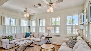 A spacious living room with a sectional sofa, a coffee table, and a round gold accent piece. There are ceiling fans, large windows with shutters, and a floor lamp. - Gulf Front Pool + Bikes! South of 30A, Steps to Beach, between Alys & Rosemary (Seacrest Beach)