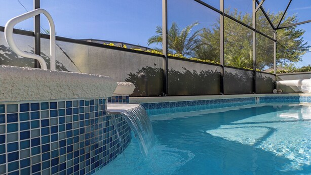 Une piscine avec un bord carrelé, une petite cascade et une vue sur les palmiers, visible depuis un espace abrité.
