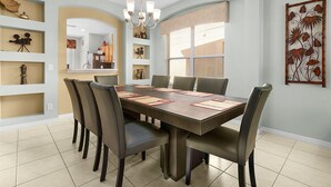 Un coin repas comprenant une grande table rectangulaire, six chaises et un lustre. Des étagères intégrées ornent les murs, et une fenêtre est équipée de stores.