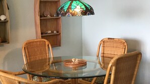 A dining area with a glass table surrounded by wicker chairs, a stained glass pendant light, and a wooden shelf with decorative items. - Panoramic Ocean and Sunset View in Poipu – Enjoy Lofty Haven Cross-Breezes! (Koloa)
