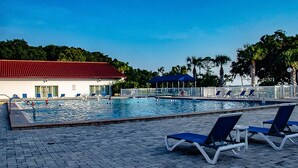 A swimming pool with lounge chairs and a covered area, surrounded by palm trees and a building with a red roof.