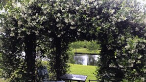 A garden table and chairs under a flowering arbor with a pond in the background.
