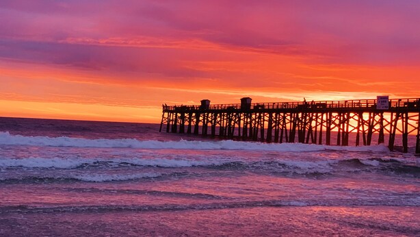 A pier extending into the ocean at sunset with waves crashing onto the shore.