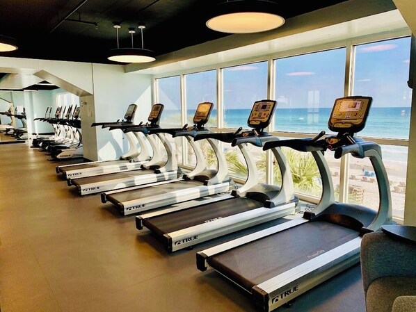 A row of treadmills with digital displays, positioned in a gym with large windows overlooking the ocean.