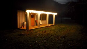 Una pequeña estructura de madera iluminada, con un porche, posiblemente un cobertizo o una cabaña pequeña, ubicada en un área cubierta de pasto con una colina al fondo.