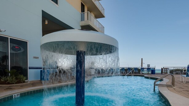 A pool with a central water feature and a building with balconies in the background. - Beachfront King Suite/Big Balcony! (Panama City Beach)