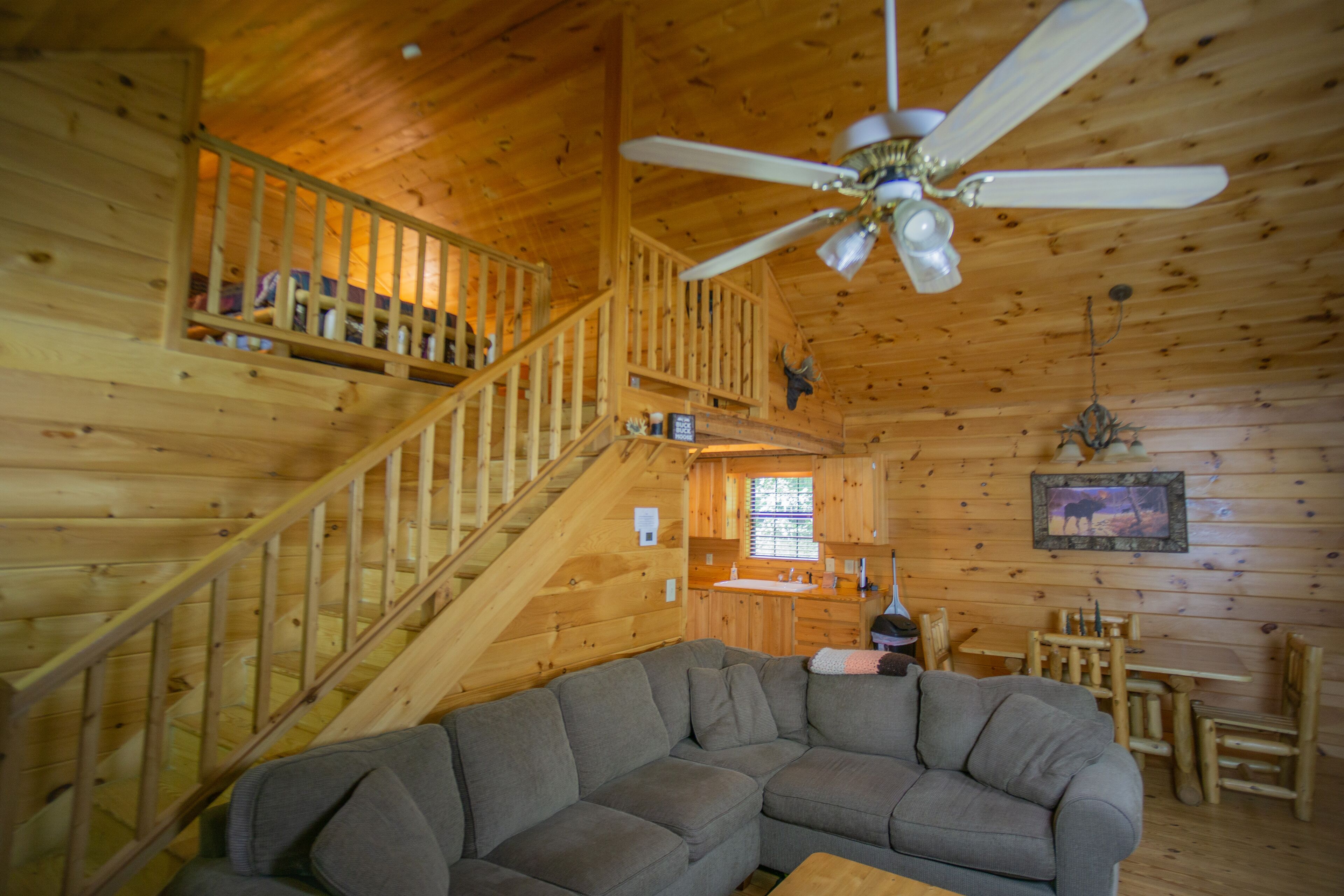 A wooden cabin interior with a staircase, a gray sectional sofa, a dining table with chairs, and a ceiling fan.