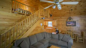 A wooden cabin interior with a staircase, a gray sectional sofa, a dining table with chairs, and a ceiling fan.