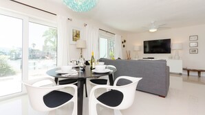 Ein modernes Wohnzimmer mit Essbereich, einem grauen Sofa, weißen Stühlen, einem Fernseher und einem Balkon mit Blick.