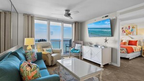 A spacious living room with a large window offering a view of the ocean, a ceiling fan, a flat-screen TV, a white cabinet, and a sofa with colorful cushions. - 16th floor 2 bedroom Beachfront at Pelican. Stunning View, 3 Pools, Beach Chairs (Destin)