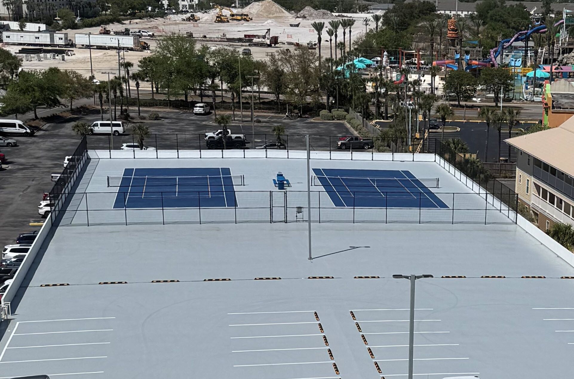 An outdoor tennis court with two courts, surrounded by a fence, with a parking lot and buildings in the background.