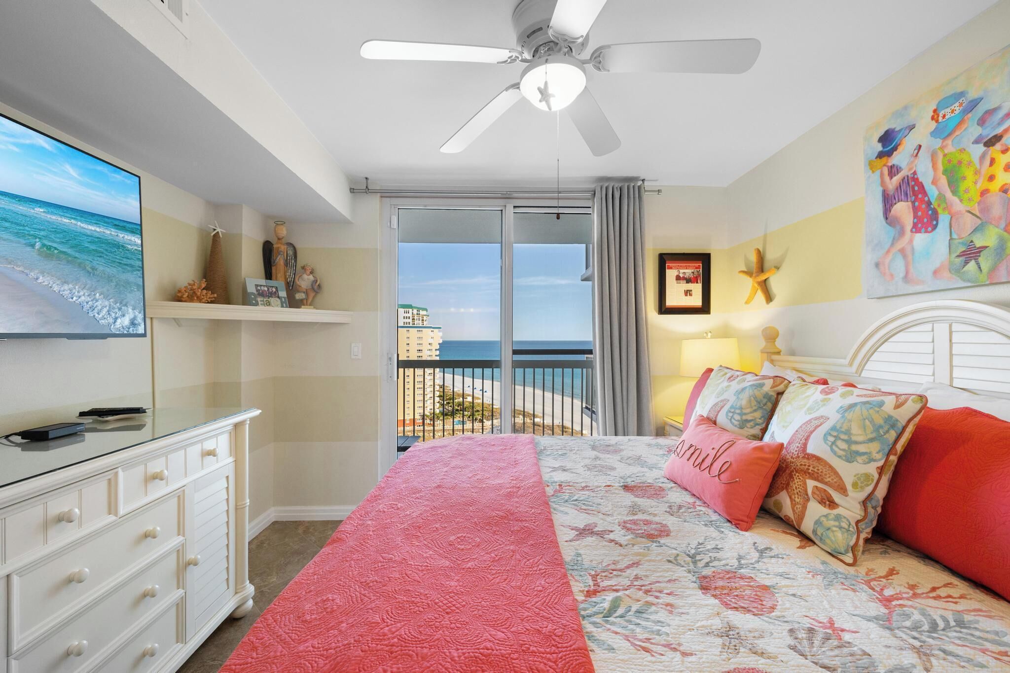 A bedroom with a bed, a TV, a ceiling fan, a balcony with a view of the ocean, and a painting on the wall.