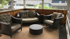 Une véranda couverte, meublée de meubles en osier, avec un pouf rond et une vue sur les maisons à travers de grandes fenêtres.