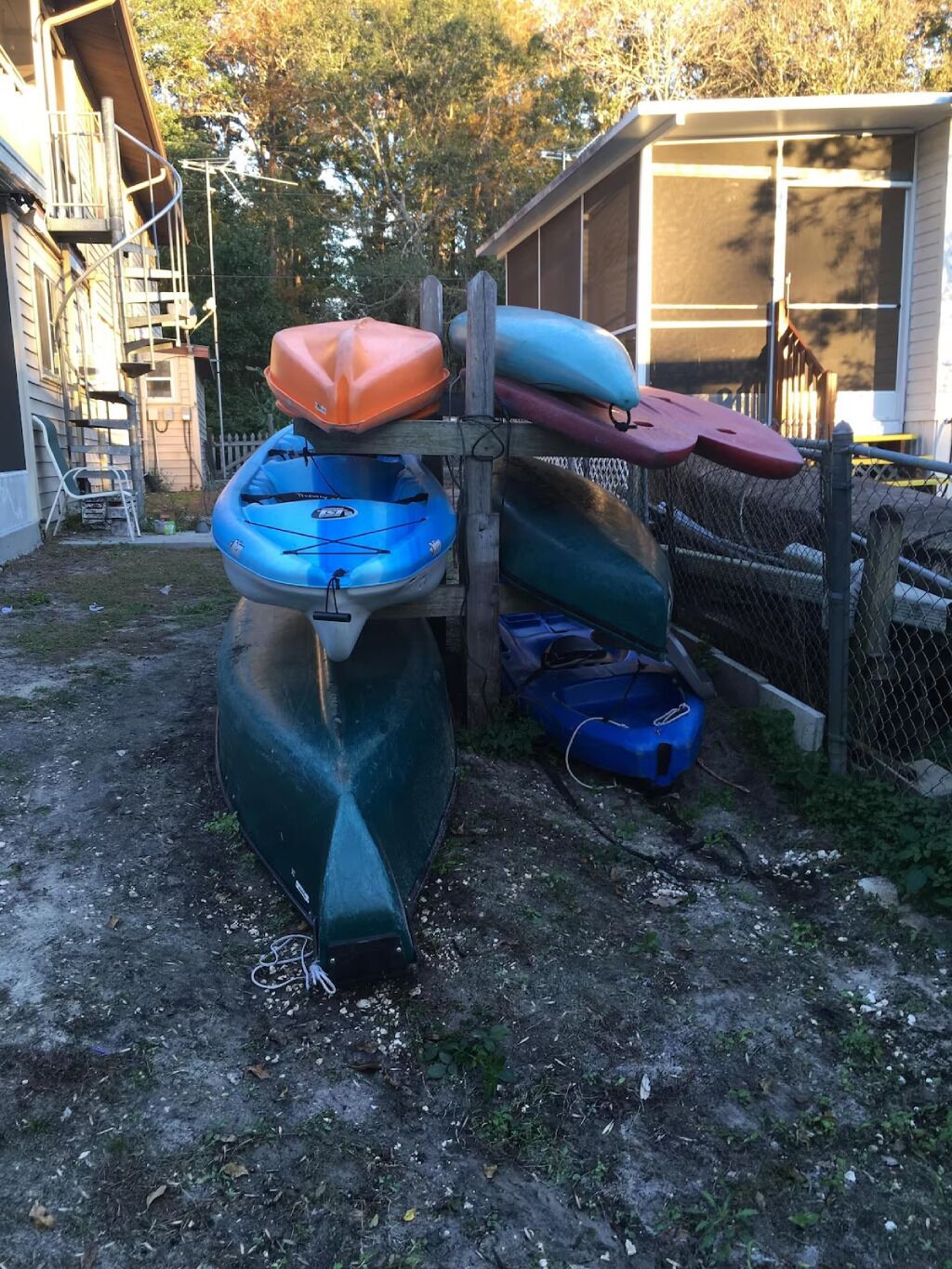 A collection of kayaks stored outdoors, a wooden fence, and a building in the background.