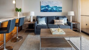 A modern living room with a dark blue sofa, two side tables with lamps, a wooden coffee table with a tray, and a large abstract painting on the wall. - COMPLETELY REMODELED Sugar Beach Oceanfront Penthouse Sunset & Ocean View (Kihei)