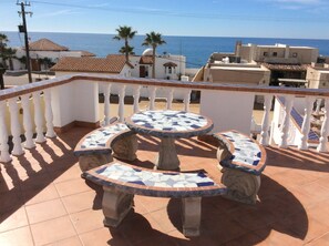Une terrasse avec une table et des chaises carrelées, une balustrade blanche et une vue sur la mer et les bâtiments.