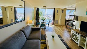 Un salon moderne avec un canapé, une table basse et un meuble pour la télévision. Il y a un balcon avec vue sur les bateaux et les palmiers.