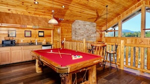 Un intérieur en bois avec une table de billard, un coin cuisine et une vue dégagée grâce à de grandes fenêtres.