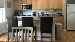 Una cocina con armarios de madera, un microondas, una estufa y un refrigerador. Hay dos sillas y un bote de basura.