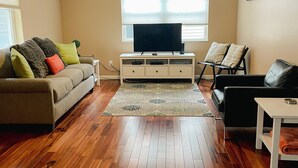 A living room with a sofa, a black leather couch, a TV on a stand, wooden flooring, and a window with blinds. - Downtown Traverse City Condo. Great Location. New King Bed  (Traverse City)