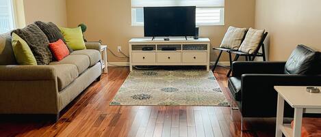 A living room with a sofa, a black leather couch, a TV on a stand, wooden flooring, and a window with blinds.