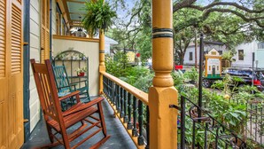 Une chaise berçante en bois sur un balcon doté d’une balustrade, donnant sur une rue bordée de voitures garées et d’immeubles.
