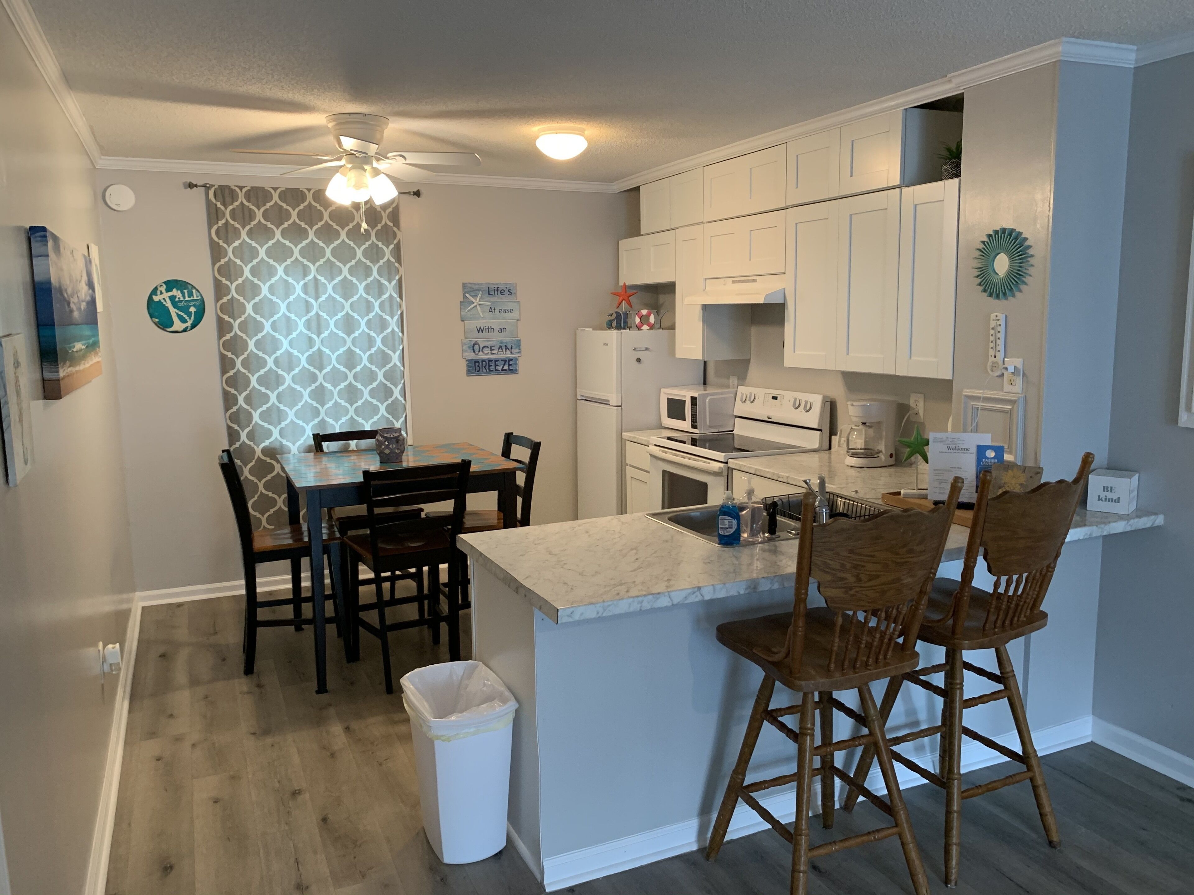 A compact kitchen with white cabinets, a microwave, a stove, and a coffee maker. There is a dining table with chairs and a trash can.