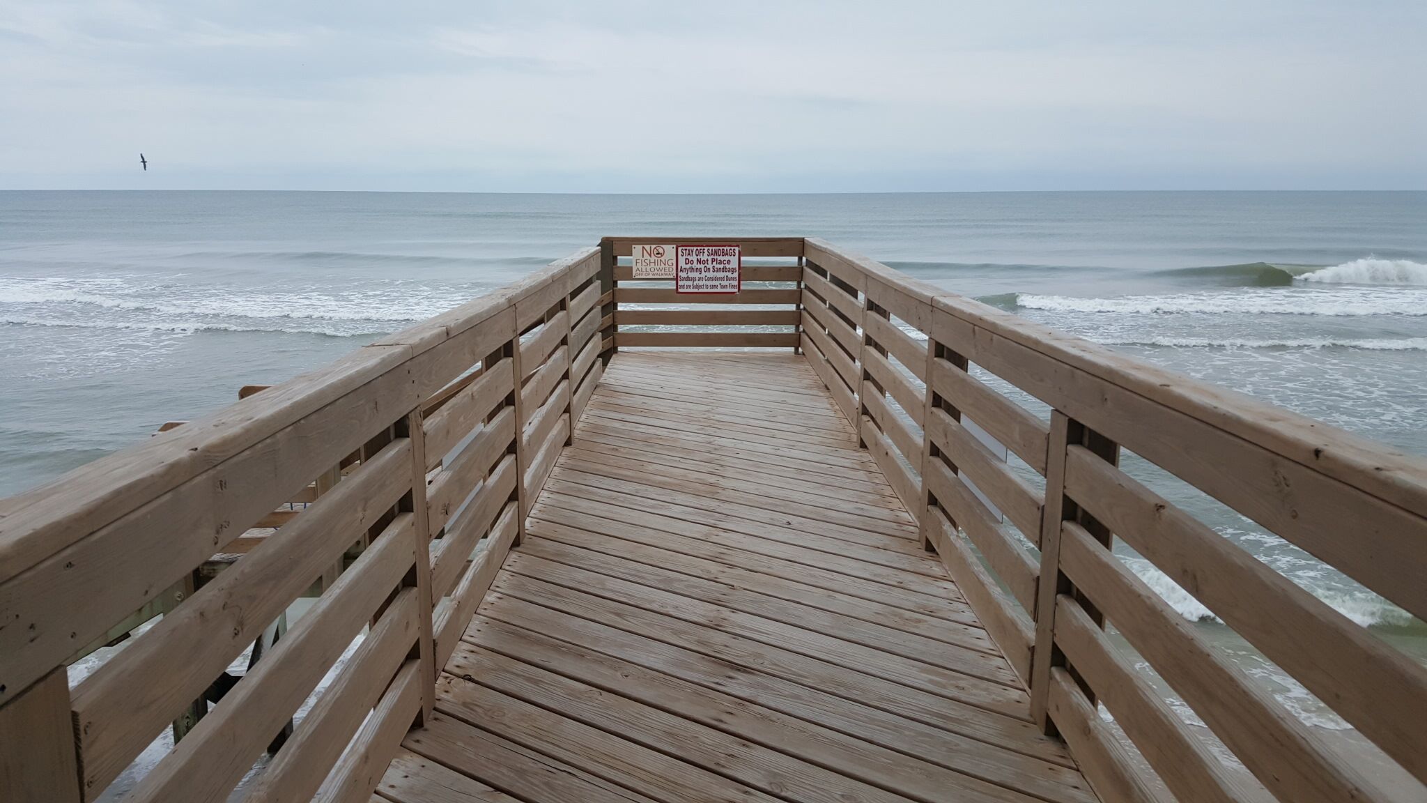 A wooden pier leading to the ocean with a sign prohibiting fishing and sandbag use.