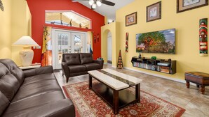 A living room with a brown leather sofa, a wooden coffee table, a red wall, a ceiling fan, and a bookshelf with various items. - Hakuna Ma'Cottage - Minecraft Room, Southern Pool! (Kissimmee)