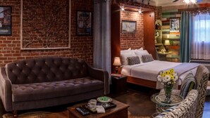A hotel room with a brick wall, a tufted sofa, a bed with a white bedspread, a patterned armchair, and a coffee table with remote controls.