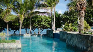 A clear blue pool surrounded by palm trees and a stone wall. - Beach Resort  Walk-Out Patio with St. John Views (St Thomas)